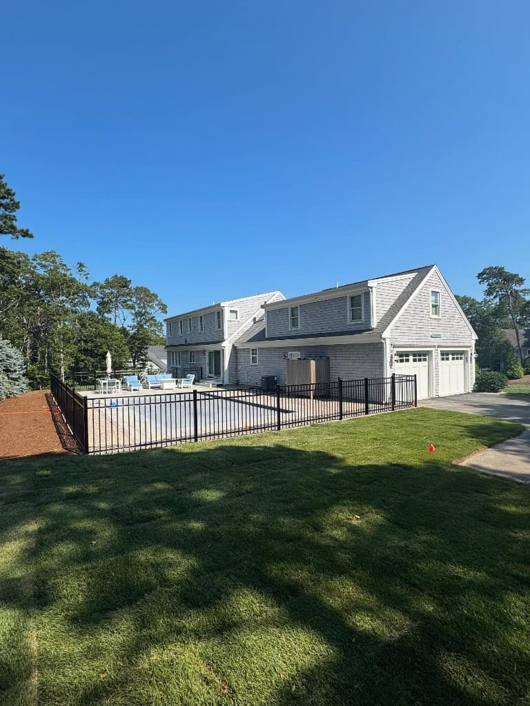 Aerial view of grey shingle Cape-style home with black metal pool fence enclosing the pool and manicured lawn — South Shore Massachusetts