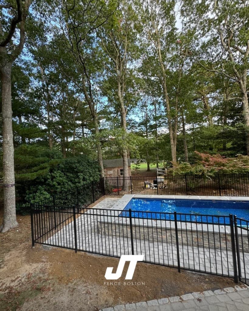 Black metal pool fence around rectangular pool in a wooded backyard with stone patio — secure barrier, natural setting